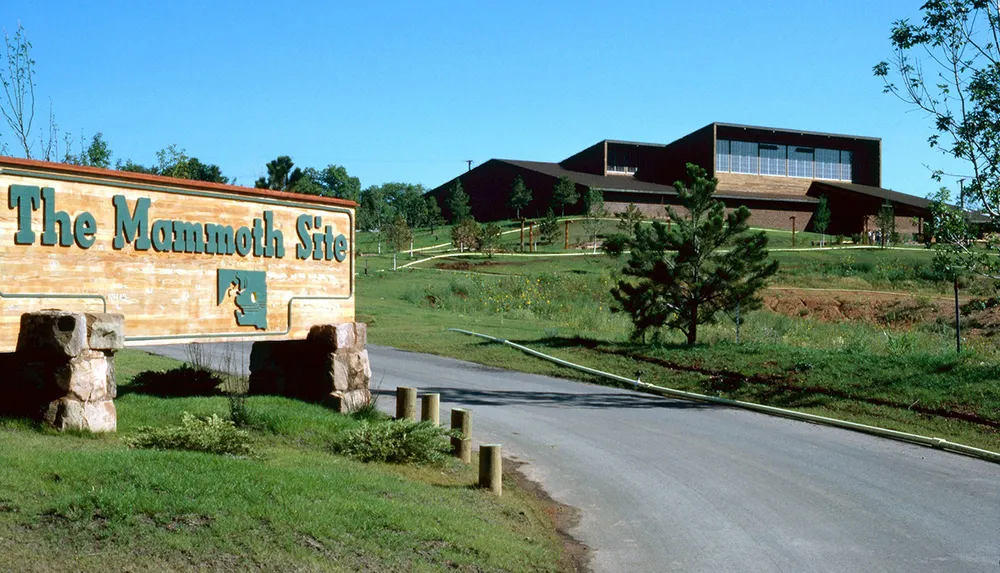 This image depicts the entrance to The Mammoth Site with a large wooden sign featuring a mammoth silhouette in front of a building on a hill with a clear blue sky above