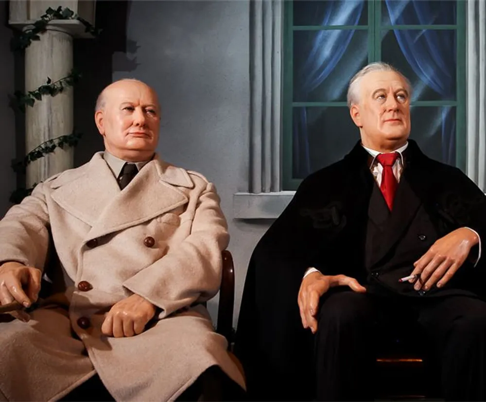 Two lifelike wax figures resembling historical figures from the mid-20th century are seated next to each other giving the impression of a pensive conversation