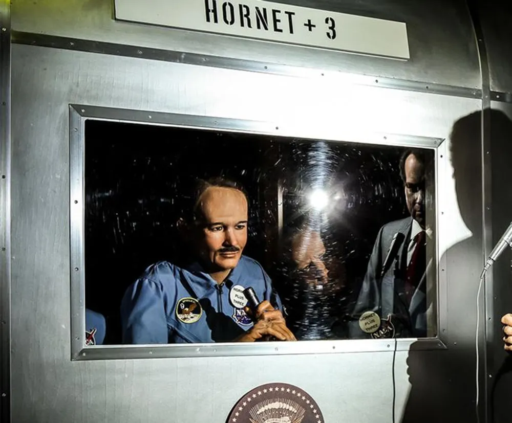 The image shows a person in a NASA jumpsuit inside a quarantine facility looking out through a small window possibly after a space mission with the sign HORNET  3 overhead