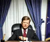 A wax figure resembling a historical figure is seated at a desk with an American flag to the right and blue curtains in the background