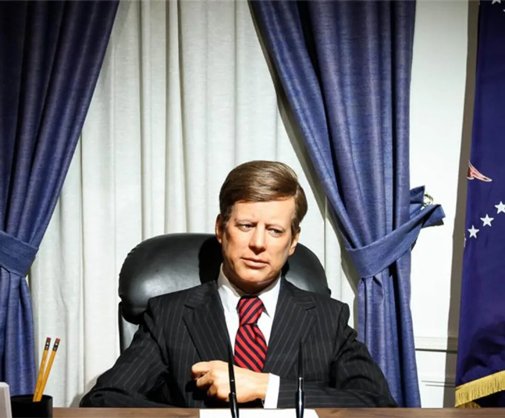 A wax figure resembling a historical figure is seated at a desk with an American flag to the right and blue curtains in the background