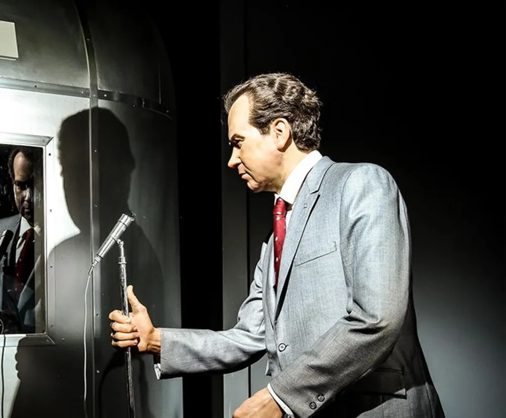 The image shows a lifelike figure of a man in a gray suit and red tie speaking into a microphone with a reflective surface behind him displaying his and the microphones reflection