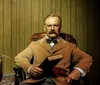 A wax figure resembling a historical figure is seated at a desk with an American flag to the right and blue curtains in the background