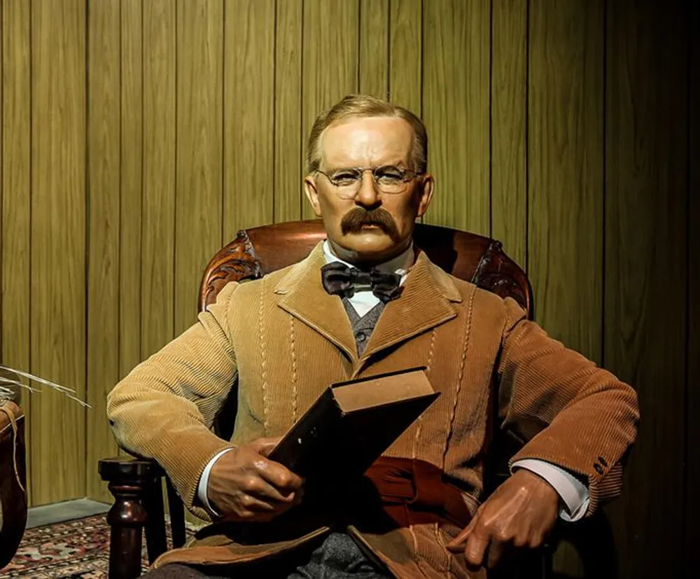 The image shows a lifelike wax figure of a seated man with a mustache wearing glasses a bow tie and a corduroy suit holding a book and giving an impression of intellectuality or scholarship