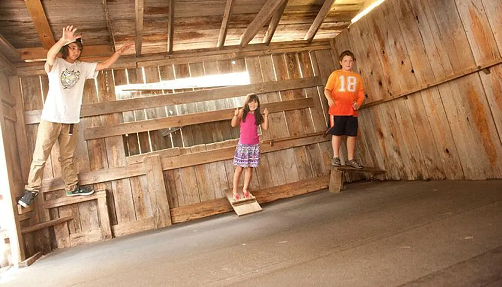 Three individuals appear to defy gravity on tilted surfaces inside what seems to be a slanted wooden room creating an optical illusion