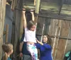 A young girl hangs from a horizontal bar with a smile while another girl supports her and onlookers gather in a rustic wooden interior