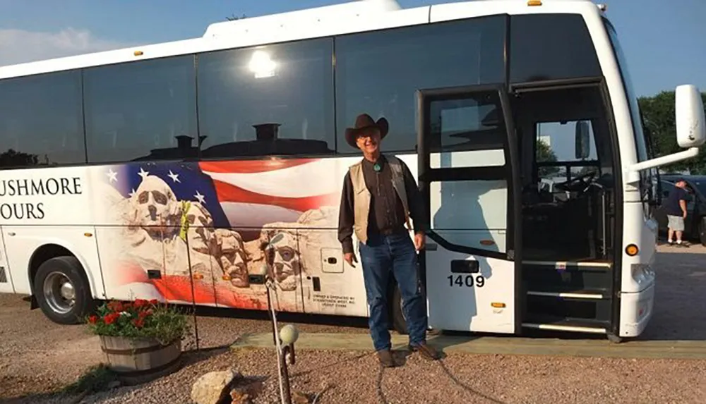 A person in a cowboy hat stands next to a tour bus adorned with an American flag and an image of Mount Rushmore