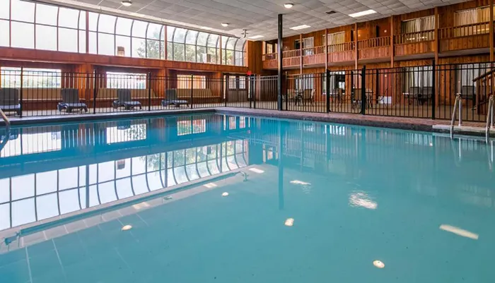 The image shows an indoor swimming pool with a serene blue water surface surrounded by multiple lounging chairs and enclosed by a building structure that features large windows and a second-level balcony with rooms all under a wooden ceiling