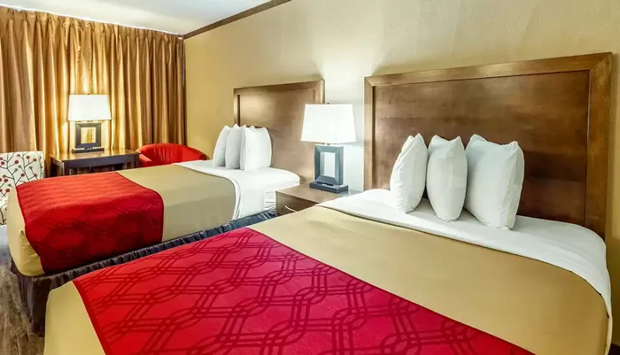 The image displays a neatly arranged hotel room with two beds featuring red and beige linens flanked by end tables with lamps and a chair in the corner