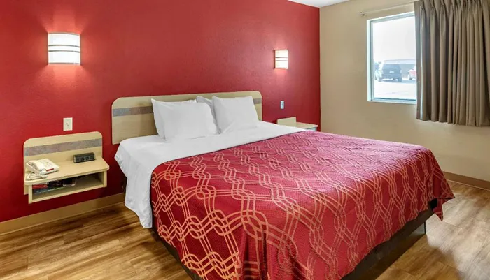 The image shows a neatly made bed with a red patterned bedspread in a room with a bold red wall on one side and a window looking out to a parking lot