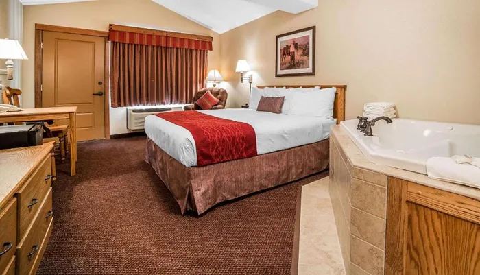 The image shows a well-appointed hotel room with a large bed featuring white and red linens accompanied by a spacious in-room whirlpool tub set against a backdrop of warm tones and classic furnishings