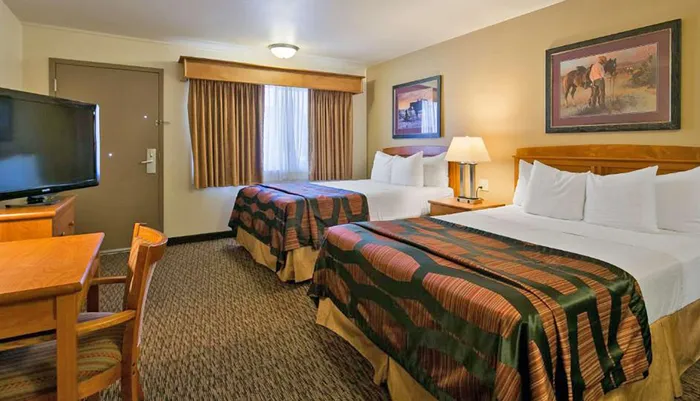 This is an image of a functional hotel room with two beds a diningwork table and a flat-screen TV decorated in a neutral color palette with a Western-themed artwork above one bed