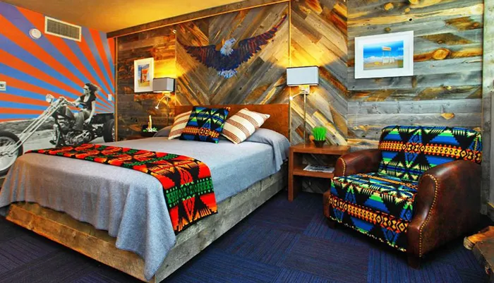 The image depicts a vibrantly decorated room with a mix of rustic wood paneling a bold eagle mural a motorbike-themed wall art colorful patterned textiles and vintage-inspired furniture creating an eclectic and spirited interior design