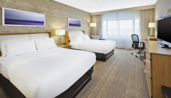The image shows a well-lit modern hotel room with two queen-sized beds a work desk a flat-screen TV and abstract art on the wall