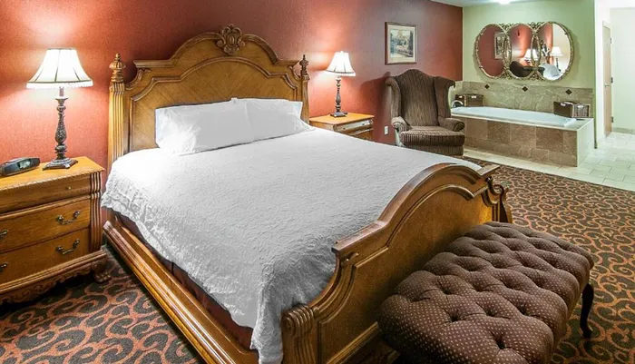 The image is of a luxurious hotel room featuring a large bed with an ornate headboard traditional furnishings and an open-concept bathtub within the room