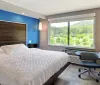 The image shows a modern hotel room with a large bed a blue accent wall a work desk with a chair and a window providing a view of green hills