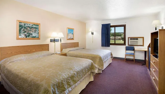 The image shows a simply furnished hotel room with two beds artwork on the walls a window with a view of the outside and a small writing desk with a chair