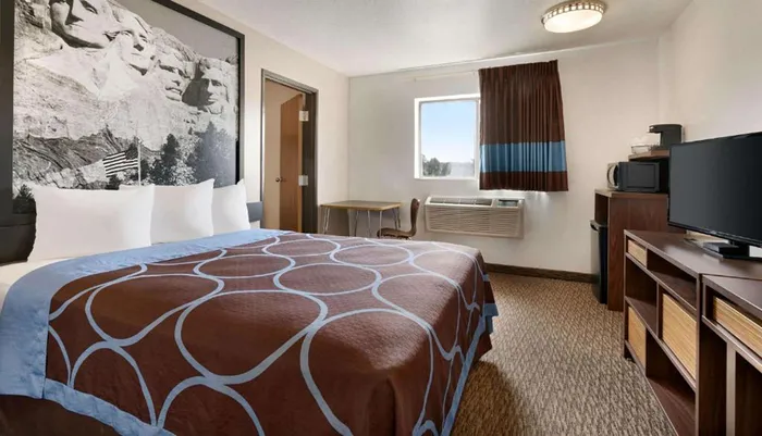 A hotel room with a large bed features a wall-size black and white photo of Mount Rushmore above the headboard a window with a brown and blue curtain basic furniture like a desk and TV stand and a patterned carpet floor