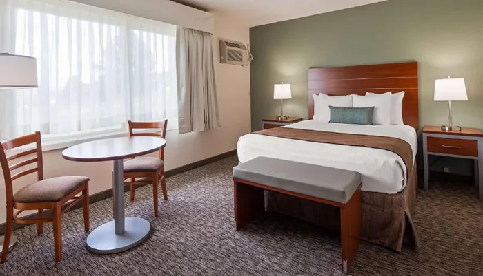 The image shows a neatly arranged hotel room with a large bed a small dining table with two chairs a bench and matching wooden furniture under ambient lighting
