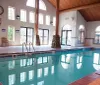 The image shows an indoor swimming pool with calm water reflecting the surrounding architecture including large windows wood beams and a vaulted ceiling