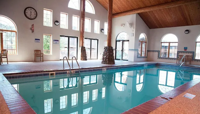 The image shows an indoor swimming pool with calm water reflecting the surrounding architecture including large windows wood beams and a vaulted ceiling