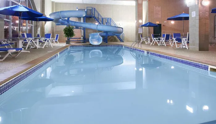 The image shows an indoor swimming pool with a blue water slide and multiple lounge chairs under umbrellas