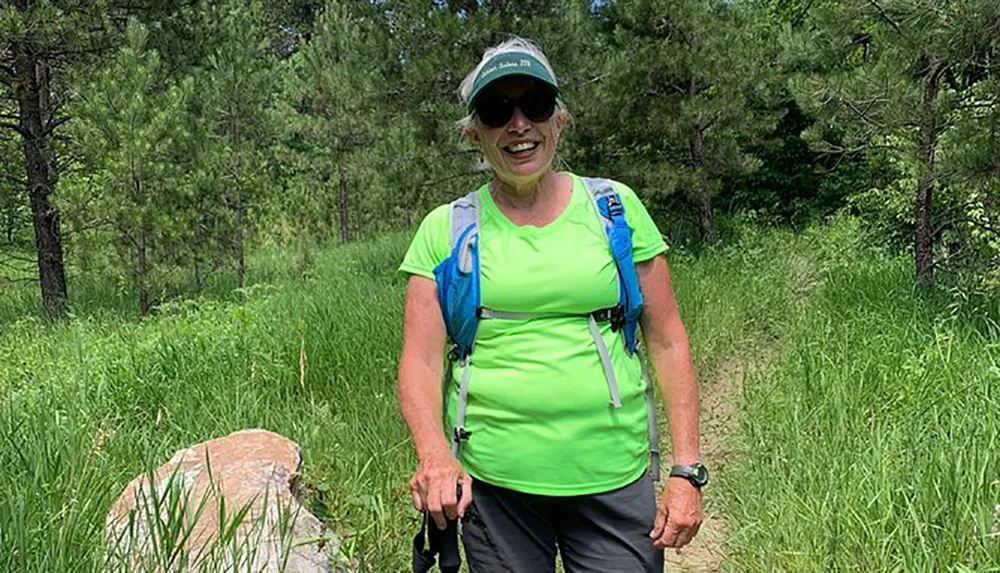 A smiling person with sunglasses and a hat is hiking on a grassy trail with a hydration pack and a walking stick surrounded by greenery and trees