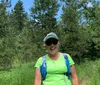 A smiling person wearing a bright green shirt sunglasses and a cap stands with a blue backpack on a grassy path among pine trees