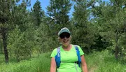 A smiling person wearing a bright green shirt, sunglasses, and a cap stands with a blue backpack on a grassy path among pine trees.