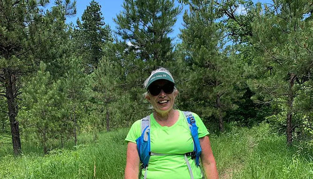 A smiling person wearing a bright green shirt sunglasses and a cap stands with a blue backpack on a grassy path among pine trees