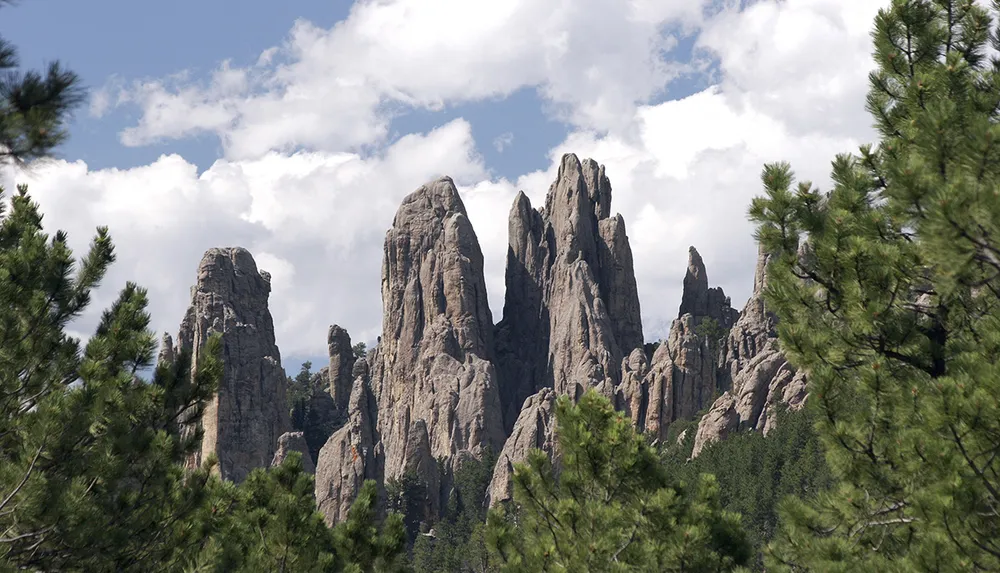 The image showcases a series of rugged towering rock formations nestled among coniferous trees under a partly cloudy sky