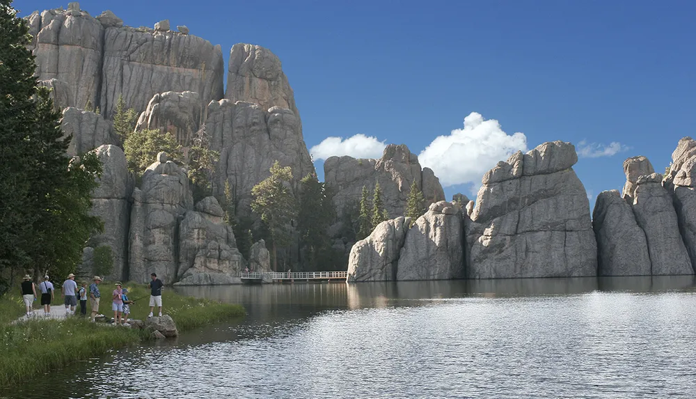 The image depicts a serene natural scene with people enjoying the outdoors by a lake that reflects towering rock formations and trees under a blue sky with wispy clouds