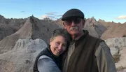 A couple is posing for a photo with a backdrop of rugged, eroded rock formations under a bright sky.