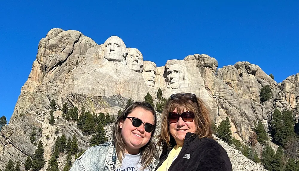 Two people are posing for a photo in front of the Mount Rushmore National Memorial which features the carved faces of four US presidents