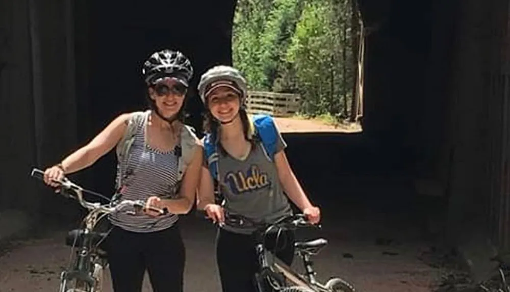 Two smiling people with bicycles are standing in front of a dark tunnel wearing helmets and casual clothing for a bike ride