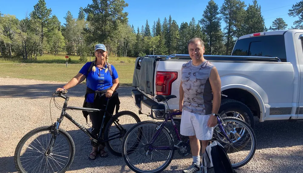 Two people are posing with their bicycles next to a pickup truck on a sunny day possibly preparing for or returning from a cycling adventure in a natural setting