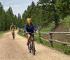 Two people are enjoying a bike ride along a gravel path lined with a wooden fence amidst green trees