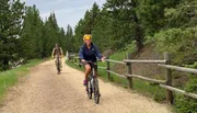 Two people are enjoying a bike ride along a gravel path lined with a wooden fence amidst green trees.