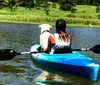 Two people are smiling and enjoying a kayak trip on a tranquil lake with a backdrop of a forested hillside partially covered in snow