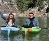 Two people are smiling and enjoying a kayak trip on a tranquil lake with a backdrop of a forested hillside partially covered in snow