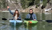 Two people are smiling and enjoying a kayak trip on a tranquil lake with a backdrop of a forested hillside partially covered in snow.