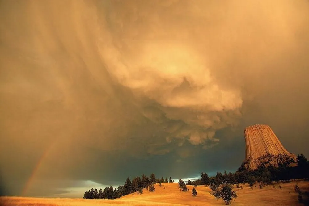 A towering cumulus cloud looms over the distinct flat-topped Devils Tower National Monument with a faint rainbow appearing on the left against a dramatic golden-lit sky