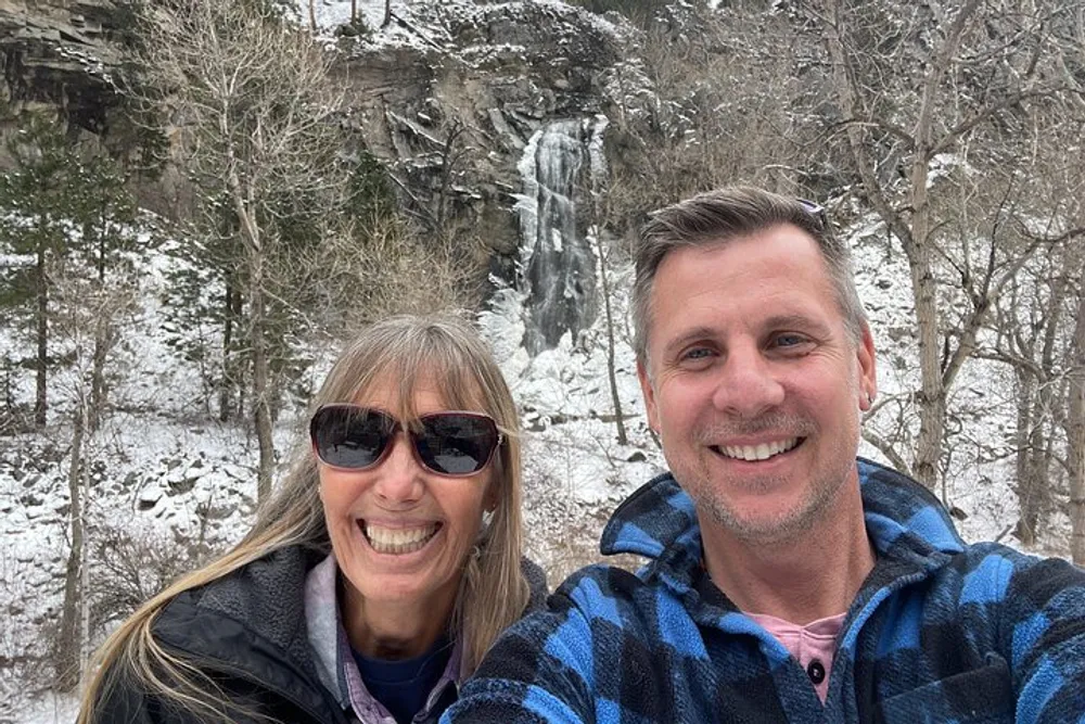 A smiling man and woman are taking a selfie with a snowy partially frozen waterfall and bare trees in the background