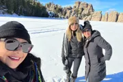 Three people are smiling for a selfie in a snowy landscape with distinctive rocky formations in the background.