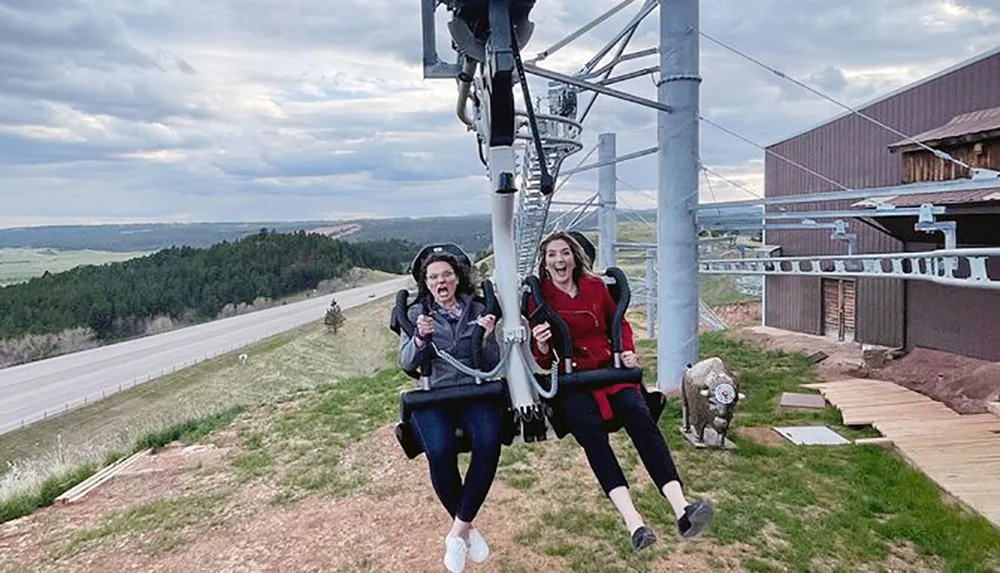 Two people are enjoying a zip line ride with one appearing thrilled and the other a bit scared as they descend near a structure with a scenic backdrop