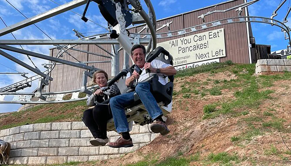 Two people are smiling and enjoying a ride on an alpine coaster near a sign that advertises All You Can Eat Pancakes