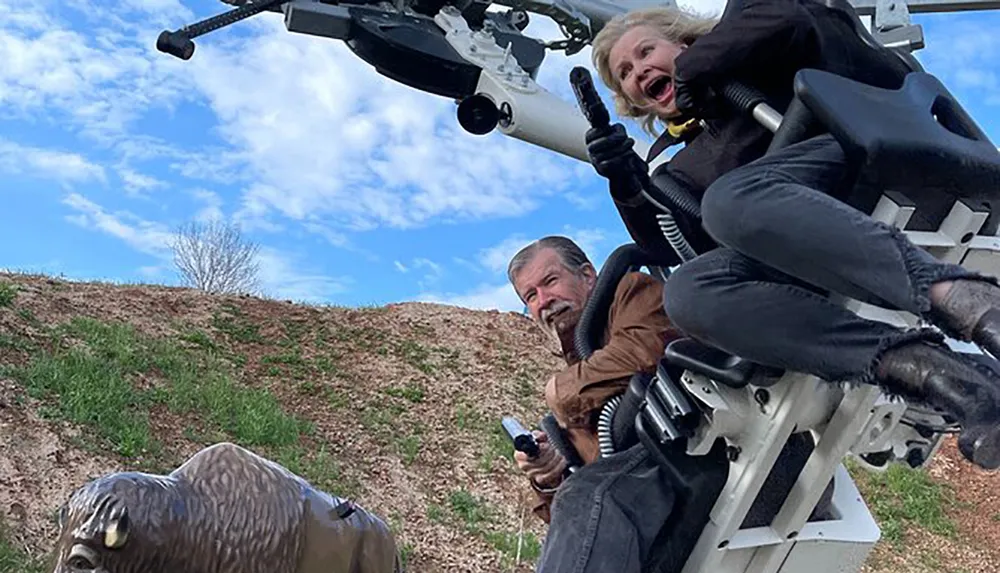Two people are strapped into a dynamic flight simulator ride with one person screaming in excitement and the other looking focused against a backdrop of a clear blue sky and grassy hill