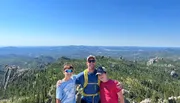 Three happy individuals are posing for a photo amidst scenic, forest-covered hills and rocky terrain under a clear blue sky.