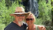 A smiling couple poses in front of a waterfall surrounded by green foliage.