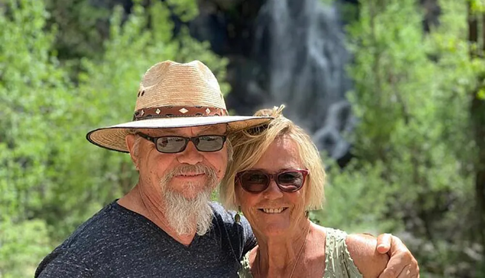 A smiling couple poses in front of a waterfall surrounded by green foliage